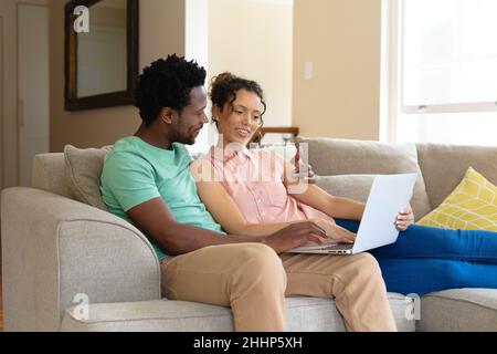 Lächelndes junges biracial Paar mit Laptop zusammen sitzen auf dem Sofa zu Hause. Menschen, Zweisamkeit und Technologie-Konzept, unverändert. Stockfoto