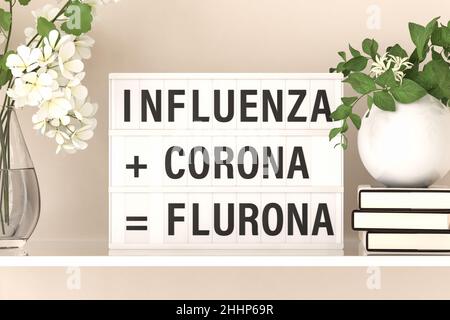 Die Worte „Influenza + Corona = Flurona“, die auf einem Leuchtkasten auf einem Regal mit Blumen und Büchern stehen. Erklärung für das neu geschaffenen Wort FL Stockfoto