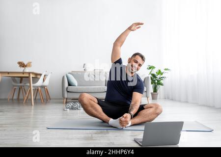 Ein glücklicher junger, muskulöser Mann aus europa sitzt auf dem Boden auf der Matte und übt sich um die Hände und schaut auf den Laptop im Wohnzimmer Stockfoto