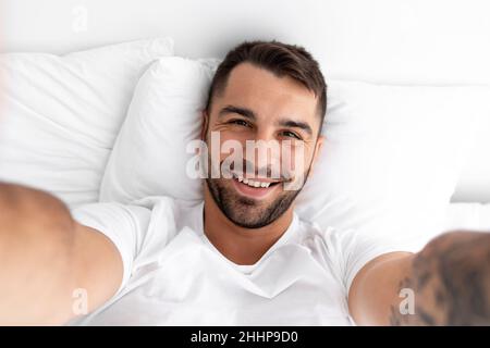 Zufrieden glücklich attraktiven jungen kaukasischen muskulösen Mann liegt auf weißem Bett und macht Selfie im Schlafzimmer, Nahaufnahme Stockfoto
