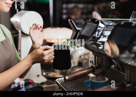 Beschnittene Ansicht des jungen Barista, der die Kanne in der Nähe der Dampfdüse der Kaffeemaschine im Café hält Stockfoto