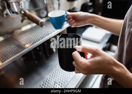 Beschnittene Ansicht des Barista, der Milchkännchen und verschwommene Tasse in der Nähe der Kaffeemaschine im Café hält Stockfoto