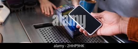 Beschnittene Ansicht des afroamerikanischen Mannes, der mit dem Mobiltelefon in der Nähe des Zahlungsterminals im Café bezahlt, Banner Stockfoto