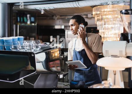 afroamerikanischer Barista, der mit dem Handy spricht und ein digitales Tablet in der Nähe der Kaffeemaschine im Café hält Stockfoto