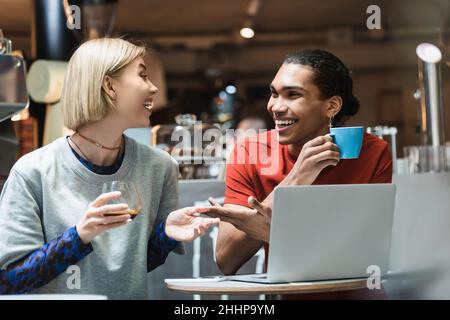 Lächelnde interracial Freiberufler reden und halten Kaffee in der Nähe von Geräten im Café Stockfoto