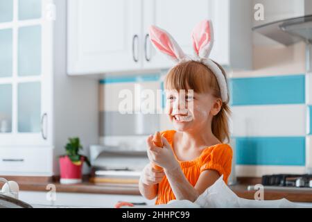 Emotionales Porträt eines fröhlichen kleinen Mädchens, das zu Ostern als Hase mit Karotte gekleidet ist, während man zu Hause in der Küche Essen kocht. Mädchen Kind Kind Spaß haben Stockfoto