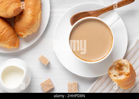 Kaffee mit Milch in einer weißen Tasse, Brötchen und brauner Zucker auf einem weißen Holztisch, Draufsicht, flach liegend. Stockfoto