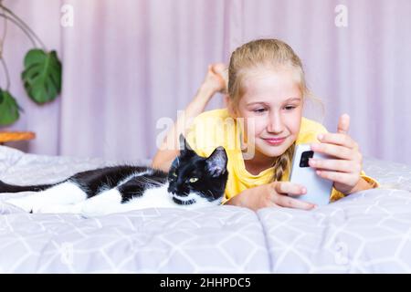 Weißes Teenager-Mädchen im gelben Sweatshirt, das auf den Bildschirm des Mobiltelefons blickt, es in den Händen hält und mit einer schwarzen und weißen Katze im Bett liegt. Kommunikation Stockfoto