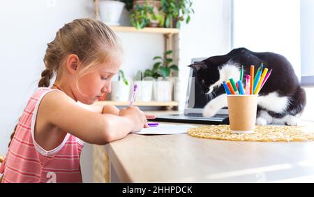 Schreiben oder Zeichnen von Kindern im Notizbuch. Eine schwarz-weiße Katze stört das Mädchen daran, ihre Hausaufgaben zu machen. Kaukasische nettes Mädchen macht Hausaufgaben zu Hause, neben Stockfoto