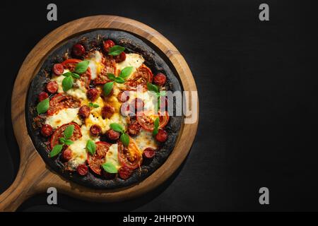 Schwarze Pizza mit Tomaten, Würstchen, Mozzarella und Basilikum. Teig mit gesundem Bambuskohlepulver. Stockfoto