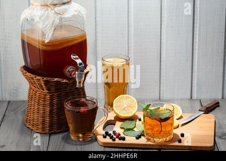 Hausgemachter Pilz-Kombucha-Drink im Glas mit Hahn und in Gläsern mit Minze, Beeren und Zitrone auf einem Holztisch. Stockfoto