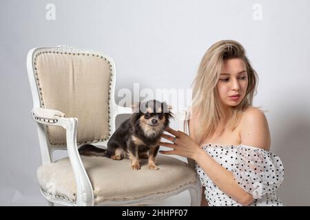Frau mit blondem Haar und weißer Haut isoliert auf grau mit kleinen schwarzen Hund im Stuhl Stockfoto