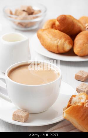 Kaffee mit Milch in einer weißen Tasse, Brötchen und braunem Zucker auf einem weißen Holztisch. Stockfoto