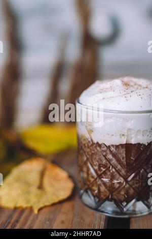 Nahaufnahme eines Glases heißen Kakaos mit Schlagsahne und Zimt, umgeben von Herbstblättern Stockfoto
