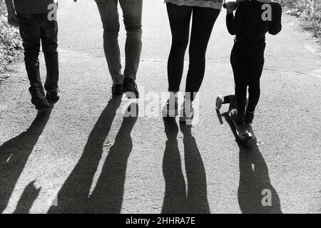 Silhouetten junger Menschen im Hintergrund. Junge Familie Vater, Mutter und zwei Kinder gehen den Weg im Park in den Strahlen der untergehenden Sonne Stockfoto