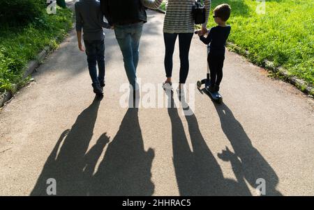 Silhouetten junger Menschen im Hintergrund. Junge Familie Vater, Mutter und zwei Kinder gehen den Weg im Park in den Strahlen der untergehenden Sonne Stockfoto