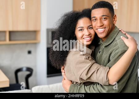 Nahaufnahme eines glücklichen multirassischen Paares, das vor verschwommenem Hintergrund in der Küche steht. Glückliche Besitzer einer neuen Wohnung lächeln und umarmen vor der Kamera Stockfoto