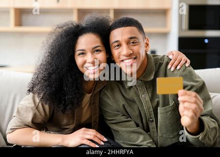 Online-Shopping. Glückliches junges Paar, das vor Freude lächelt, während es mit Kreditkarte auf dem Sofa sitzt und auf die Kamera schaut. Glücklicher Biracial Mann zeigt Plastikkreditkarte Stockfoto