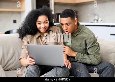 Birazialfrau und ihr Mann schauten auf den Laptop und freuten sich über den Erfolg. Fröhlicher Kerl bekam eine Beförderung bei der Arbeit, gewann die Lotterie, kaufte ein Urlaubsticket Stockfoto