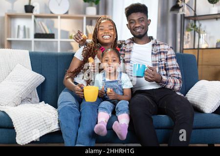 Junge Eltern und Tochter lächeln und blicken auf die Kamera, während sie sich auf dem Sofa mit heißem Tee und frischen Croissants ausruhen. Porträt einer glücklichen afroamerikanischen Familie. Stockfoto