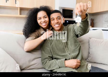 Umzugskonzept. Glückliches multirassisches Paar, das Schlüssel aus der neuen Wohnung zeigt, während es in Umarmungen im gemütlichen Wohnzimmer sitzt. Fröhliche Besitzer in einem neuen Hotel Stockfoto