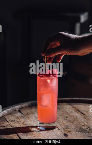 Hohes rosafarbenes Cocktailgetränk mit drei Himbeeren auf Cocktailpickel, auf Holzfass Stockfoto