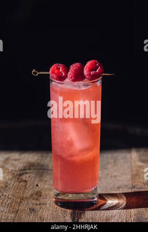 Hohes rosafarbenes Cocktailgetränk mit drei Himbeeren auf Cocktailpickel, auf Holzfass Stockfoto