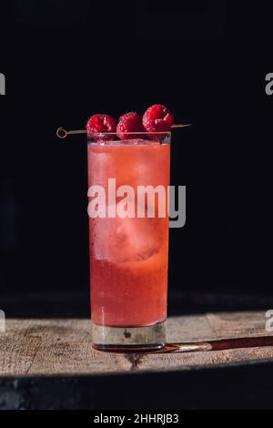 Hohes rosafarbenes Cocktailgetränk mit drei Himbeeren auf Cocktailpickel, auf Holzfass Stockfoto