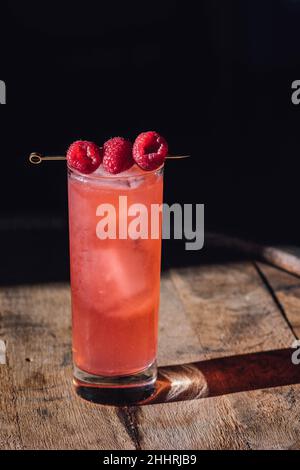 Hohes rosafarbenes Cocktailgetränk mit drei Himbeeren auf Cocktailpickel, auf Holzfass Stockfoto