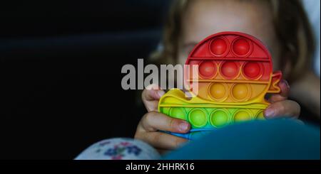 Kleines Kind spielt mit Regenbogen Pop-it Fidget Gadget, das ein beliebtes Silikon Anti-Stress-Spielzeug ist Stockfoto