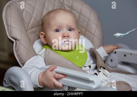 Porträt eines Babys, das zum ersten Mal mit einem Löffel auf einem Hochstuhl isst. Stockfoto