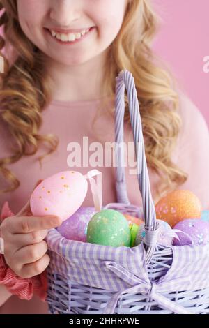 Nahaufnahme eines lächelnden, stylischen Kindes in rosa Kleid mit Korb mit Ostereiern auf rosa Hintergrund. Stockfoto