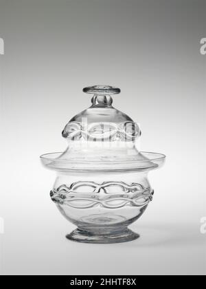 Zuckerdose 1812–27 Thomas Cains zugeschrieben die kettenartige Dekoration dieser Zuckerschale und des Känkels (69,167) wurde durch das Aufbringen zweier paralleler Glasfäden erreicht, die dann zu den miteinander verbundenen Ovalen zusammengezerreißt wurden. Diese Art der Dekoration geht auf die antike römische Zeit zurück und wurde auf venezianischem und englischem Glas aus dem 16. Jahrhundert wiederbelebt. In der zweiten Hälfte des 18. Jahrhunderts erschien es wieder auf englischem Glas. Thomas Cains, der aus Bristol, England, kam, war Inhaber beider Firmen, die diese Stücke möglicherweise hergestellt haben. Zuckerdose 7957 Stockfoto