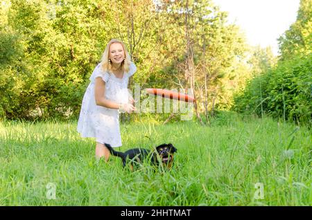 Eine blonde junge Frau in einem leichten Kleid wirft eine fliegende Scheibe, nach der ein süßer Hund rennt. Stockfoto