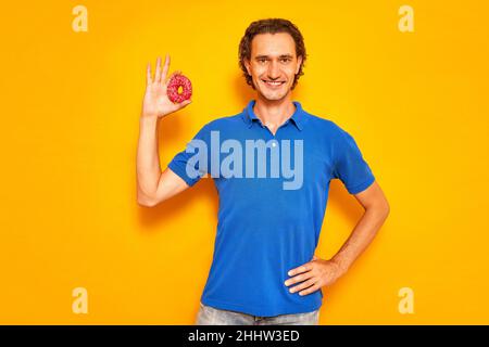 Lächelnder Mann hält Donut in der Hand in Form eines OK-Zeichens, er ist in legerer Kleidung, blauem Polo gekleidet Stockfoto