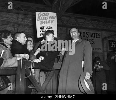 Der amerikanische Evangelist Billy Graham begrüßt die riesige Menschenmenge, die sich nach seiner Ankunft des französischen Liners Liberte versammelt hatte, um ihn am Plymouth Dock Gates zu begrüßen. Hier singt er eine Hymne, die von der siebenjährigen Giles Cooper beobachtet wird, die auf den Schrankenstrahlen sitzt. 18th. März 1955. Stockfoto