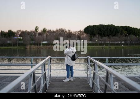 Vertikale Aufnahme einer Frau mit dicker Kapuzenjacke, die Fotos vom Steg an einem See gemacht hat Stockfoto