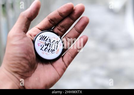 Hand eines Mannes, der einen runden Schlüsselbund mit der Aufschrift „Mixed with Love“ auf einem weichen, unscharfen Hintergrund hält Stockfoto