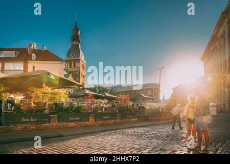 Riga, Lettland. Junge Paar Touristen Menschen Zu Fuß Auf Dome Square Im Sonnigen Sommertag. Dome Cathedral Auf Dem Hintergrund Bei Sonnenuntergang. Stockfoto