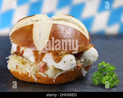 Kalbswurst mit süßem Senf und Sauerkraut in einem Brezelbrötchen, serviert auf einem schwarzen Schieferteller mit Petersilie Stockfoto