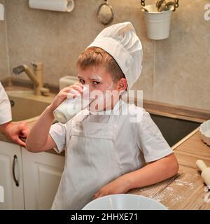 Ein Junge in der Kleidung des Küchenchefs trinkt Milch aus einem Glas, während er in der Küche einen Apfelkuchen kocht Stockfoto