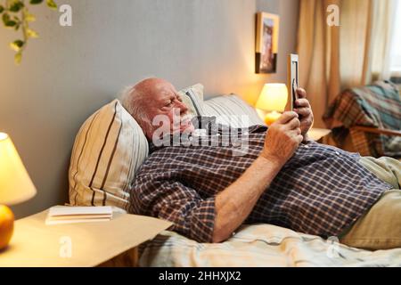 Alter Mann in Casualwear, der auf dem Bett liegt und sich das Bild seiner Frau ansieht, während er zu Hause bleibt und das Wochenende alleine verbringt Stockfoto