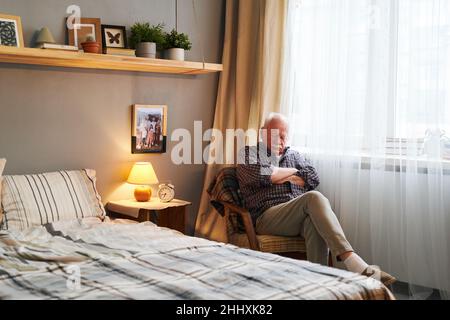 Müder älterer Mann, der in einem Sessel am Fenster sitzt und mitten am Tag vor dem Doppelbett in der häuslichen Umgebung nippt Stockfoto