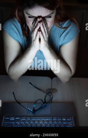 Frau arbeitet Überstunden. Ein Student, der nachts im Dunkeln an einem Laptop studiert. Das Mädchen macht sich Sorgen über Probleme bei der Arbeit und hält ihre Hände auf ihrem Gesicht. Stockfoto