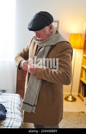 Älterer Mann knöpfte eleganten Mantel, während er im Wohnzimmer seiner Wohnung stand und sich vor dem Ausgehen anzog Stockfoto
