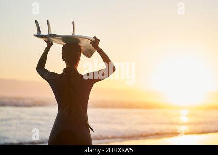 Surfen ist eine Lebensart. Rückansicht einer Surferin, die ihr Surfbrett am Strand über ihren Kopf trägt. Stockfoto