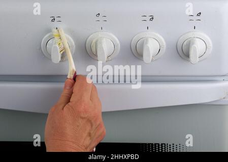 Weibliche Hand wäscht die Wasserhähne des weißen Gasofens mit einer Zahnbürste. Eine ungewöhnliche Art der Hausreinigung Stockfoto