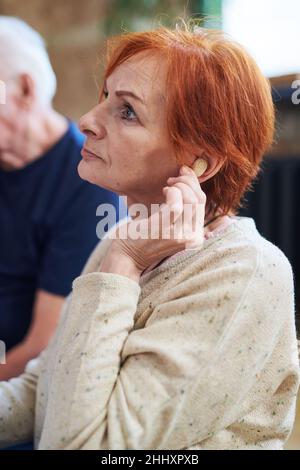 Eine ältere Frau im Pullover berührt ihr Ohr mit einem Hörgerät, während sie im Fitnessstudio gegen einen älteren Mann in einem blauen T-Shirt sitzt Stockfoto
