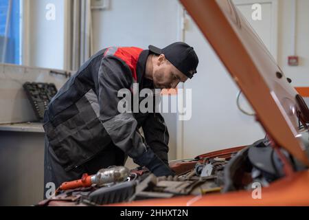 Junger Mann in Arbeitskleidung, der sich mit dem Motor über die offene Motorhaube beugt, während er sie vor der Reparatur in der Werkstatt überprüft Stockfoto