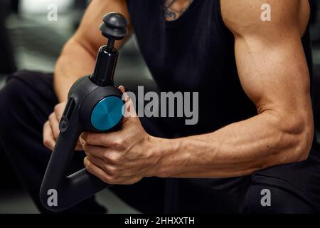 Ein junger Mann benutzt und genießt die Schlagmassagepistole, um sich zu entspannen und die Schmerzen im Körper und in den Muskeln im Fitnessstudio zu lindern. Stockfoto